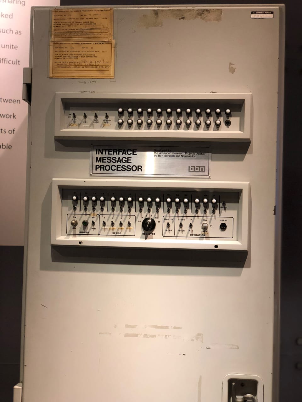 A tall grey metal cabinet with two panels of small switches and lights. A sign on it says Interface Message Processor.
