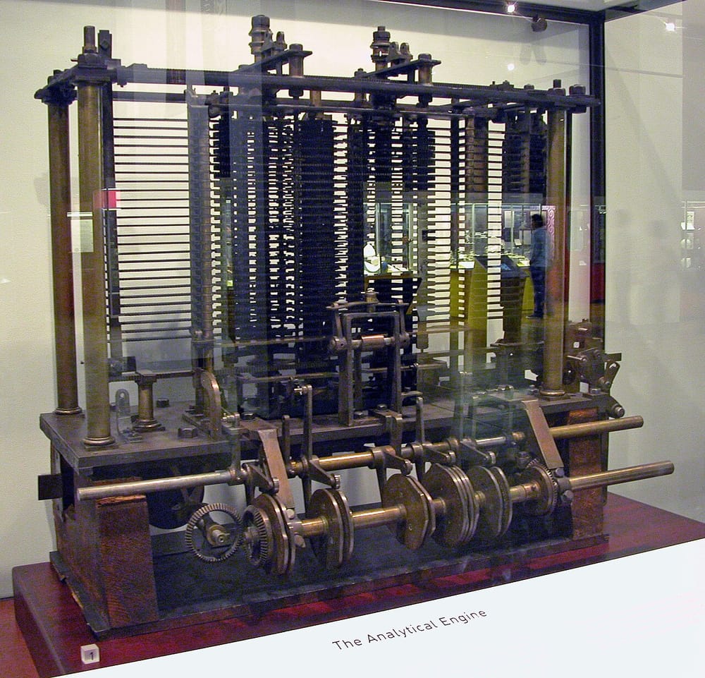 The trial piece of the Analytical Engine in a glass case: a metal frame packed with stacked gears, with rods and wheels at the front.