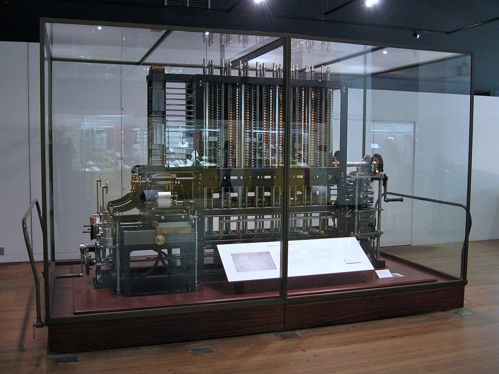 Difference Engine No. 2 in a glass case at the Science Museum, London: a long machine of tall metal columns full of gears, with a crank handle at one end.