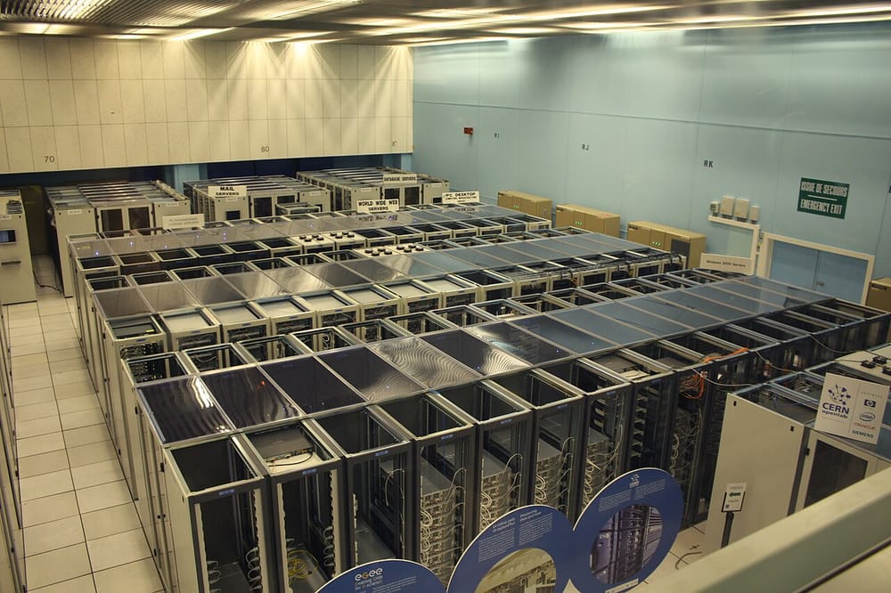 A large, bright room seen from above, with rows of tall metal cabinets (racks) full of computer equipment and cables. Signs above the rows name groups of servers.