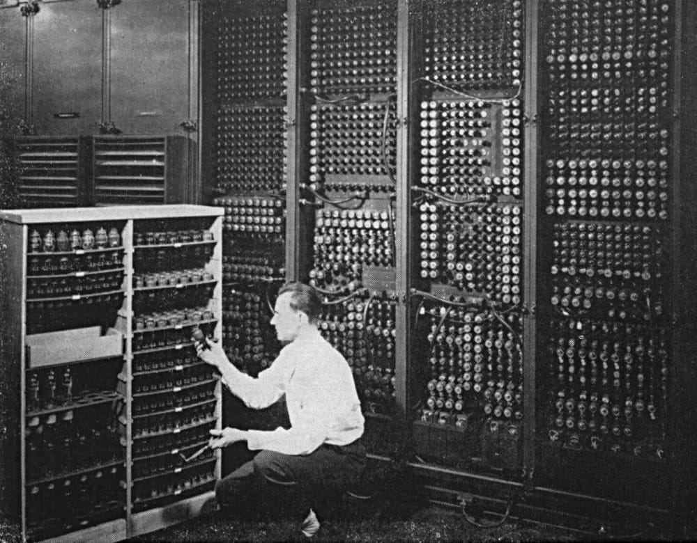 A man kneels in front of ENIAC holding a tube, with rows of vacuum tubes behind him.