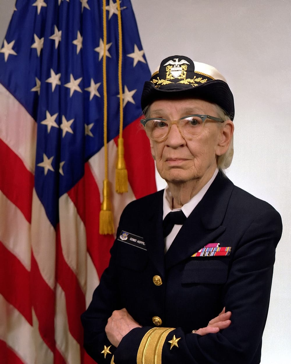 Grace Hopper, an older woman with glasses, in a dark US Navy uniform and cap, arms folded, in front of a US flag.