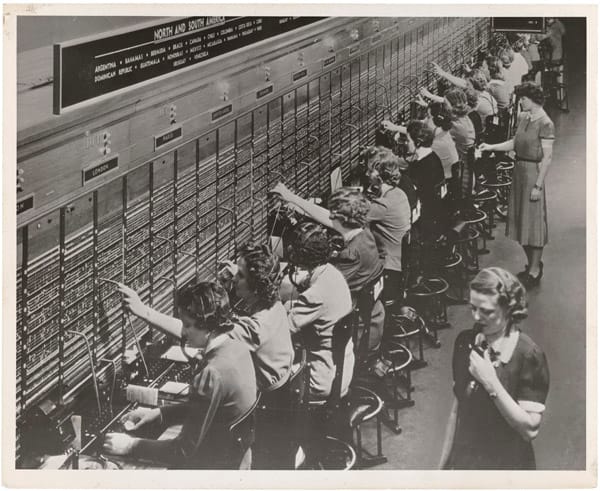 1940s: women operators with headsets at a long switchboard, with cords plugged into its sockets.
