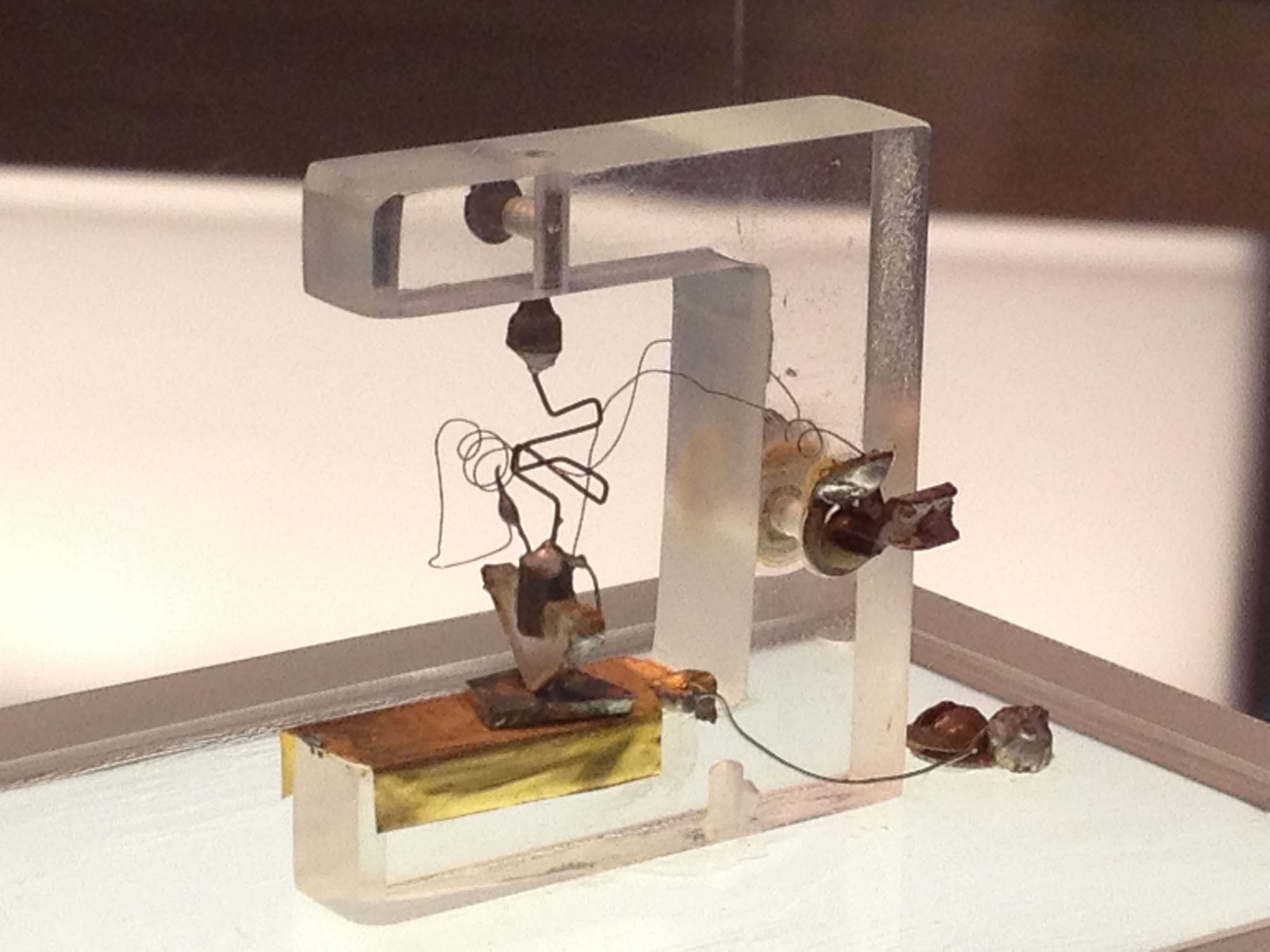 The first transistor from 1947, in a museum case. A clear plastic frame shaped like a hook holds a bent wire. At the bottom, a small plastic wedge shaped like a triangle points down onto a small, flat, dark piece of germanium, which sits on a gold-coloured metal block.